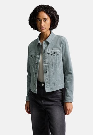 Giacca di jeans azzurro chiaro con colletto, due tasche sul petto, bottoni frontali e design corto. Indossata sopra una maglietta bianca e jeans neri.