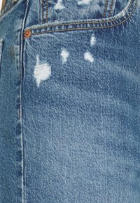 Gros plan sur un jean en denim bleu avec des zones usées près de la poche avant et des coutures orange visibles.