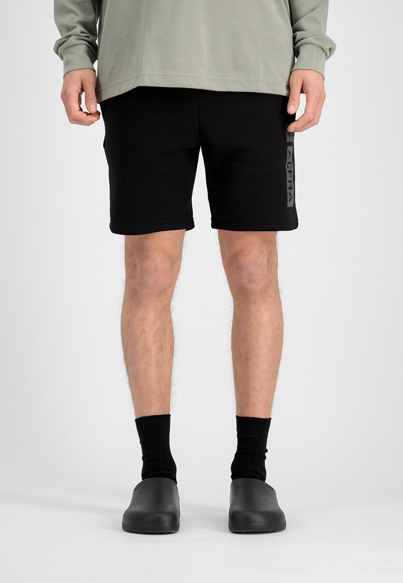 Shorts noirs avec un logo sur le côté, associés à des chaussettes noires et des chaussures gris à enfiler. La tenue présente une coupe décontractée et une texture lisse.