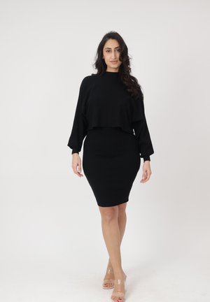 Frau mit langem dunklem Haar, die ein schwarzes langärmliges Oberteil und einen eng anliegenden schwarzen Rock trägt, geht vorwärts vor einem einfarbigen hellen Hintergrund.