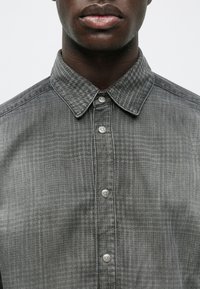 Prim-plan al unei persoane care poartă o cămașă cu carouri gri, cu nasturi din metal și guler ascuțit.