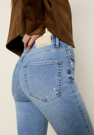 Des jeans en denim bleu clair présentent une coupe ajustée, des poches arrière, un léger effet usé et une étiquette beige de marque au niveau de la taille.