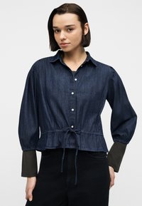 Γυναίκα με κοντά σκούρα μαλλιά που φοράει σκούρο τζιν πουκάμισο με κουμπιά και ρυθμιζόμενο κορδόνι στη μέση, καθώς και μαύρο παντελόνι, μπροστά σε μονόχρωμο φόντο.