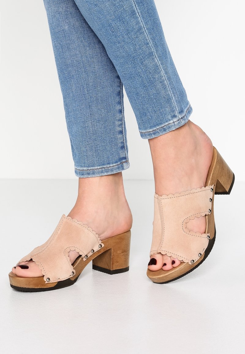 Mules beiges en daim à bout ouvert avec des bords festonnés, ornées de clous métalliques, montées sur un talon épais en bois brun.