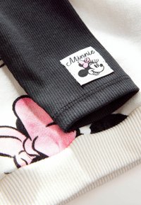 Manche noire côtelée avec un patch Minnie Mouse, présentant des accents roses. La partie inférieure est en tissu blanc avec un motif Minnie Mouse en rose et noir.