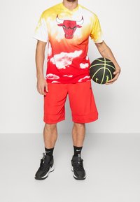 Koszulka z gradientem czerwonym i żółtym z grafiką byka, zestawiona z czerwonymi szortami. Czarna sportowa obuwie z białymi akcentami i piłka do koszykówki.