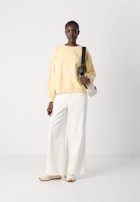 Ljust gult oversized sweatshirt, vita vida byxor och vita sneakers med beige sulor; modellen håller en liten grå väska med svart rem.