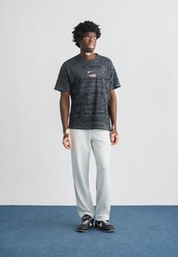 Μαύρο μπλουζάκι Nike με σχέδιο και μικρό κόκκινο και λευκό λογότυπο, σε συνδυασμό με ανοιχτό γκρι φόρμα και μαύρα αθλητικά παπούτσια.