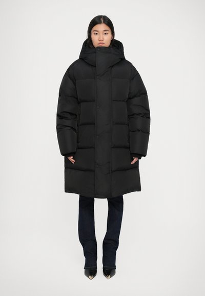 Doudoune noire avec capuche, présentant un design matelassé, une coupe oversize, des boutons-pression et des poignets élastiques, associée à un jean sombre et des chaussures pointues.