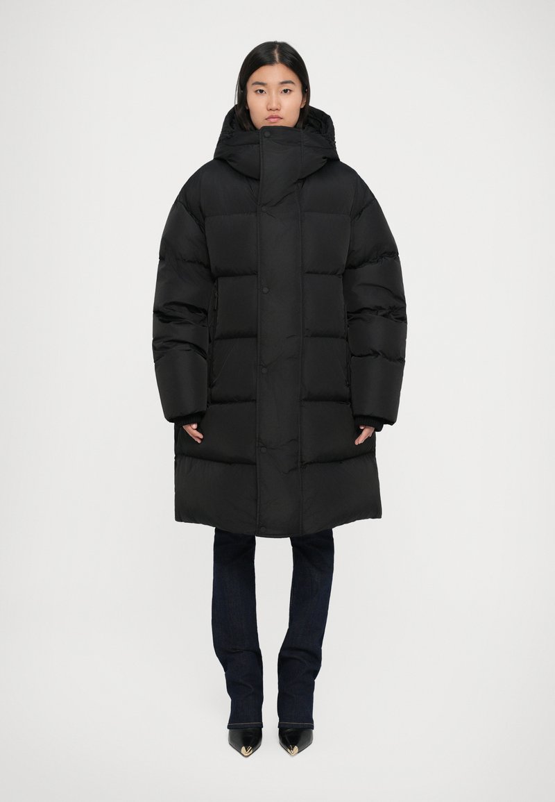 Giacca a vento nera con cappuccio, caratterizzata da un design trapuntato, vestibilità oversize, bottoni a pressione e polsini elasticizzati, abbinata a jeans scuri e scarpe con punta.
