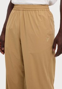 Mann in beigefarbenen, elastischen Hose mit Seitentaschen und einem kleinen "7"-Logo neben der rechten Tasche, stehend vor einem weißen Hintergrund.