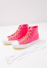 Des baskets montantes rose vif avec des semelles en caoutchouc blanches et un détail étoile jaune, des lacets jaunes et des lacets blancs supplémentaires à côté.