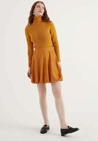 Maglione a collo alto a coste color senape abbinato a una gonna a campana strutturata dello stesso colore. Mocassini neri completano l'outfit.