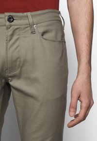 Světle hnědé legíny se zapínáním na přední knoflík, bočními kapsami a viditelnými švy. Hladká tkanina s přiléhavým střihem.