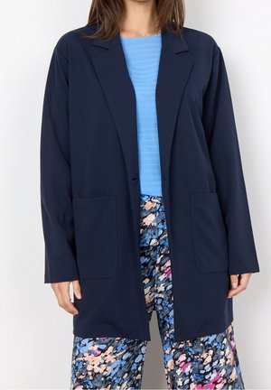 Femme portant un blazer bleu marine ouvert devant, un haut bleu clair et un pantalon à motif floral multicolore.