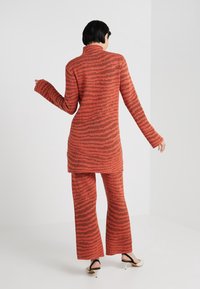 Personne aux cheveux courts et foncés portant un ensemble assorti à rayures orange et marron, composé d'un haut à manches longues et d'un pantalon, debout de dos à la caméra.