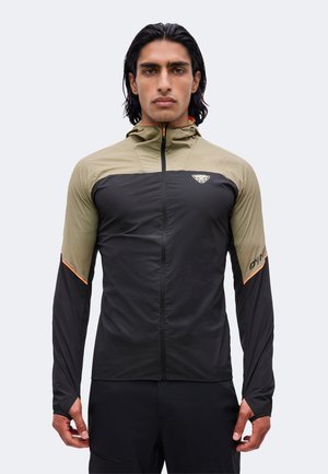 Uomo con capelli scuri indossa una giacca slim-fit nera e verde oliva con zip, fori per i pollici e un piccolo logo sul petto.