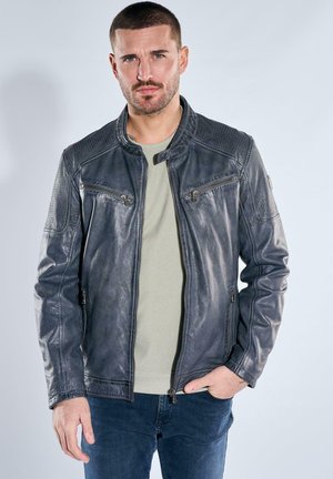 Mann mit kurzen Haaren, der eine dunkelgraue Lederjacke über einem hellgrauen Pullover und blauen Jeans trägt, stehend vor einem schlichten hellen Hintergrund.
