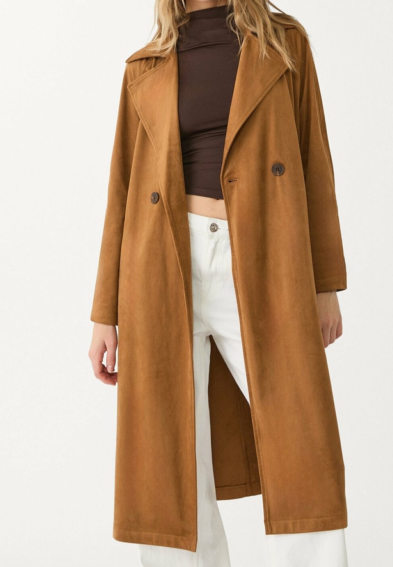 Femme portant un long manteau croisé camel brun par-dessus un col roulé court marron foncé et un pantalon blanc taille haute.