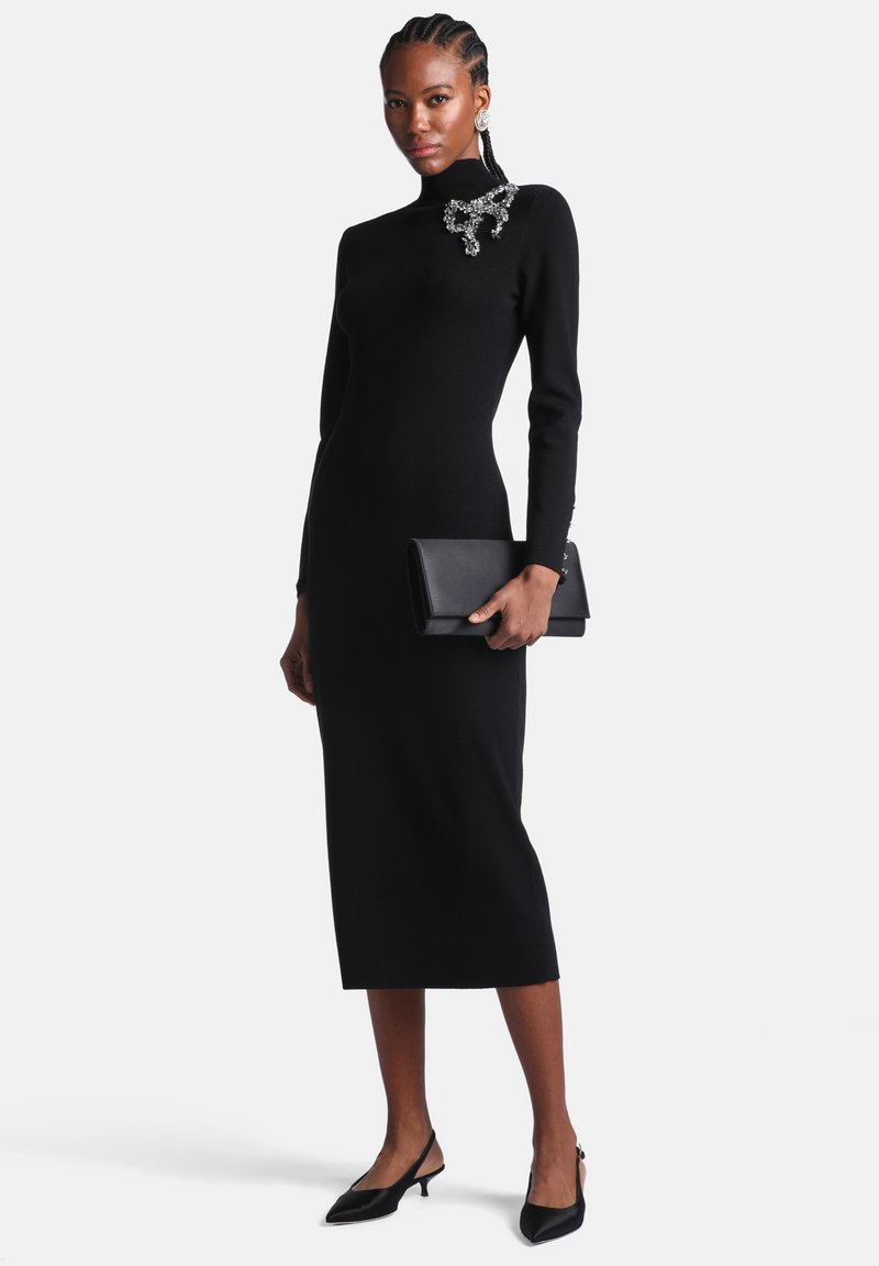 Femme portant une robe midi noire ajustée à manches longues avec une broche décorative, tenant une pochette noire et portant des escarpins slingback noirs.
