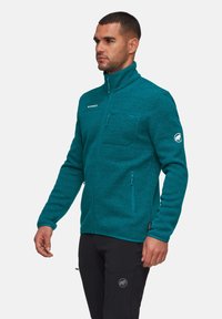 Giacca in pile teal con collo alto, tasche frontali e logo Mammut. Presenta un motivo testurizzato e chiusure a zip, indossata con pantaloni neri.