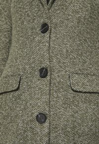 Blazer texturizado verde oliva con un patrón tejido, dos bolsillos frontales y botones negros para el cierre.