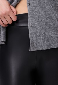 Gray knit cardigan with buttons partially covering black high-waisted leggings. The leggings have a smooth, glossy texture.