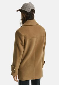 Camel-wollmantel mit lässiger Passform, breitem Reverskragen und Knopfmanschetten in Goldoptik. Kombiniert mit einer grauen Mütze mit brauem Schirm.