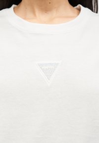 Άσπρο πουκάμισο με στρογγυλή λαιμόκοψη και μικρό τρίγωνο λογότυπο Guess διακοσμημένο με στρας στο στήθος.