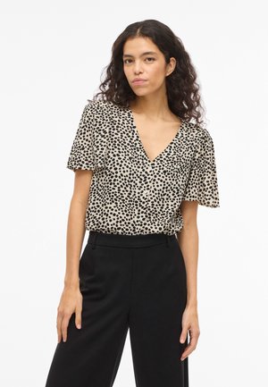 Ženska s kodrastimi lasmi, oblečena v bež kratkoročno bluzo z črnimi pikami in črne hlače z visokim pasom, na belem ozadju.