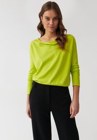 Limettengrüner, langärmeliger Pullover mit einem Bootsausschnitt, der einen lockeren Sitz und eine weiche Textur aufweist, kombiniert mit hochtaillierten schwarzen Hosen.