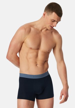 Muskelöser Mann schaut nach unten, trägt schwarze Boxer-Shorts mit grauem Bund vor weißem, einfarbigem Hintergrund.