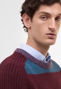 Knit sweater featuring maroon, teal, and purple sections, ribbed texture, and a crew neck layered over a light blue collared shirt.