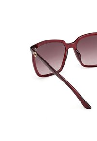 Sluneční brýle ve stylu kočičích očí z hlubokého červeného plastu, s gradientovými čočkami a zlatým akcentem na straně. Elegantní, úhlový design s zúženými ramínky.