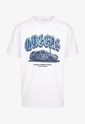 Biała, oversize'owa koszulka z niebieskim napisem "QUEENS" oraz ilustracją, zestawiona z beżowymi spodniami cargo i białymi trampkami.