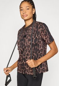 Sportinis marškinėlis su leopardų raštu rudos ir juodos spalvos, trumpomis rankovėmis ir apvalia iškirpte, ant krūtinės su siuvinėtu logotipu.