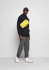 Bolso bandolera amarillo, sudadera con capucha negra, pantalones grises, zapatillas blancas. El bolso cuenta con un cierre de cremallera, una marca sutil y una textura suave.