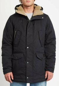 Schwarze isolierte Parka mit beigem Fleece-gefüttertem Kapuze, zwei großen Fronttaschen, einer gepolsterten Brusttasche und Druckknopfverschluss. Lässige Passform.