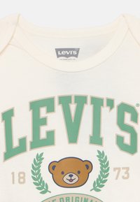 Krämfärgad Levi's skjorta med grön text, ett leende björnhuvud mellan gröna lagerkvistar, samt siffrorna 18 och 73 på vardera sida.