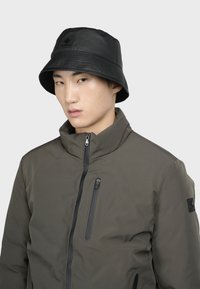 Cappello a secchiello nero con visiera testurizzata e logo. Il modello indossa una giacca verde oliva con zip e tasca sul petto con zip e patch sulla spalla.