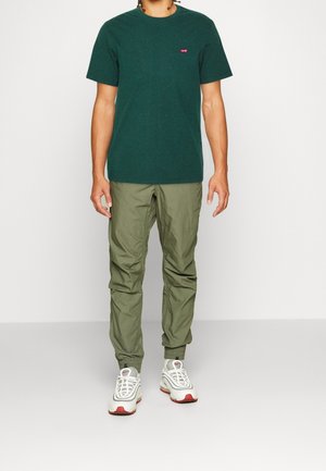 Grünes Baumwoll-T-Shirt mit Rundhalsausschnitt, kleinem roten Logo auf der Brust, kombiniert mit helloliven, schmal zulaufenden Cargo-Hosen und weißen Sneakern mit roten Akzenten.