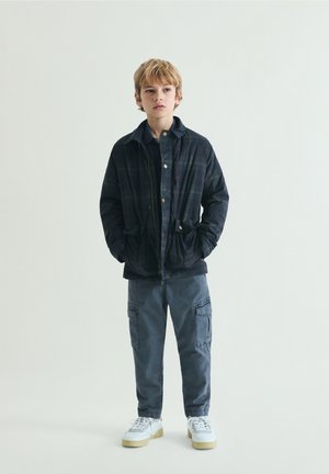 Blonder Junge steht mit den Händen in den Taschen, trägt eine dunkle Jacke, blaue Cargohosen und weiße Turnschuhe vor einem einfarbigen hellen Hintergrund.