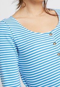 Blå och vit randig topp i strukturerad väv, med rundad halsringning och tre guldknappar framtill.