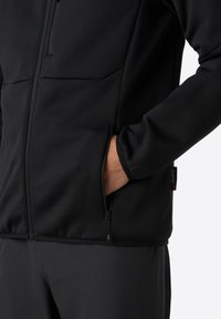 Schwarze Fleecejacke mit Reißverschluss vorne, seitlichen Taschen und weicher Textur, mit einem Etikett mit der Aufschrift "Polartec" an der Seite.
