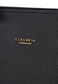 Černá kožená taška s texturou, která má pozlacené embosované logo s nápisem "CARLHEIM COPENHAGEN". Detailní záběr na materiál a branding.