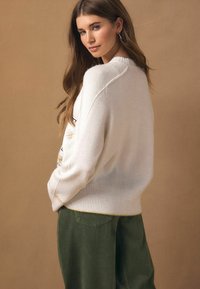 Trui van zacht witte garen met geribbelde manchetten en zoom, voorzien van meerkleurige borduursels en een ontspannen pasvorm. Olijfgroene ribfluwelen broek.