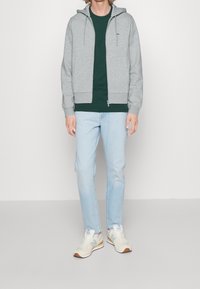 Sudadera gris con cremallera y cordones, llevada sobre una camiseta verde oscuro. Jeans de color azul claro y zapatillas beige con acentos naranjas completan el conjunto.