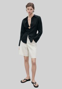 Zwarte linnen overhemd met lange mouwen, onbevouwd. Crème gestreepte shorts en zwarte teenslippers. Minimalistisch ontwerp met een relaxte pasvorm.