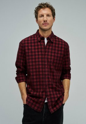 Homme aux cheveux bouclés portant une chemise à carreaux rouge et noir avec les manches retroussées et un pantalon noir, les mains dans les poches.