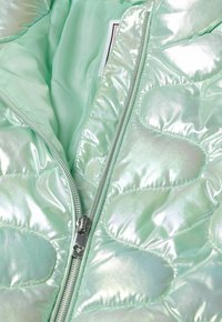 Veste matelassée irisée vert clair avec fermeture éclair argentée partiellement ouverte, révélant une doublure intérieure lisse et des motifs cousus en forme de cœur.